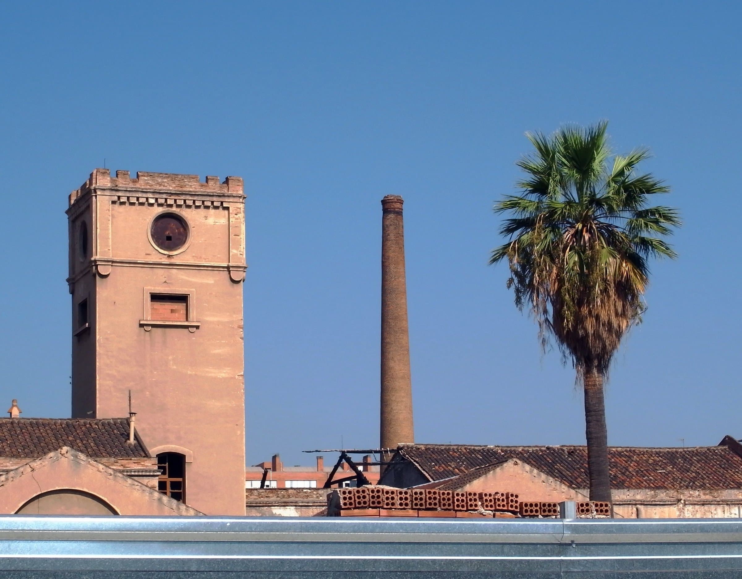 Torre de una antigua fábrica, una chimenea de ladrillo y una palmera en el carrer del Marroc, en el Poblenou de Barcelona.